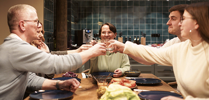 Eine glückliche Familie sitzt bei einem gemütlichen Abendessen beisammen und prostet sich mit den erhobenen Gläsern zu.